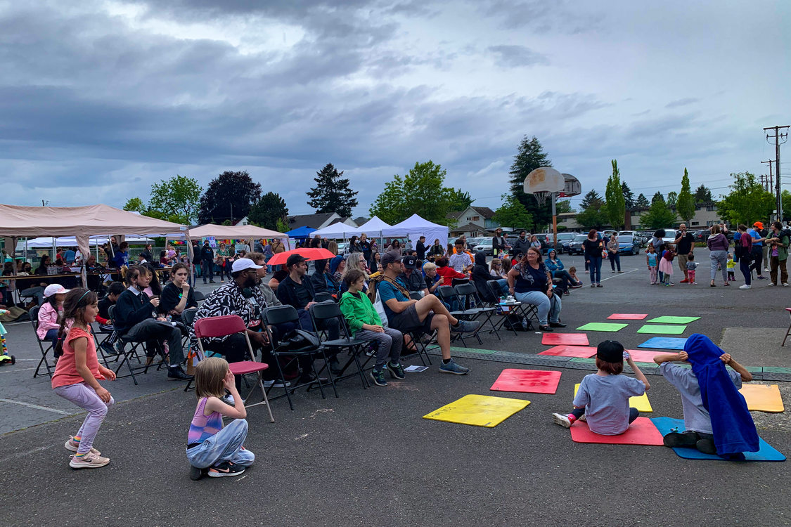 Parents and community members sit in rows of chairs set up on a school blacktop as they watch performances at Vestal Social Justice Night. Younger children sit on yoga mats on the ground and play nearby.