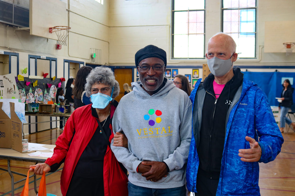 A light-skinned woman, dark-skinned man and light-skinned man attend the Vestal Social Justice Night in a gym with projects set up throughout the space