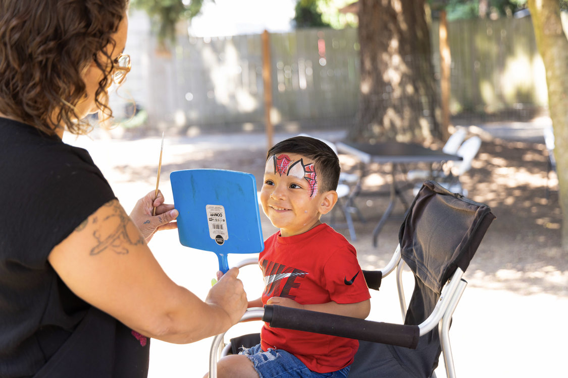 A young light brown skinned boy with face paint smiles while looking in a mirror the face painter holds up for him.