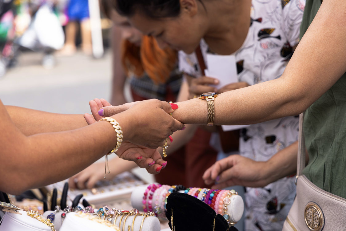 A vendor’s hand ties a bracelet onto a customer's hand above a table covered in brightly colored jewelry.