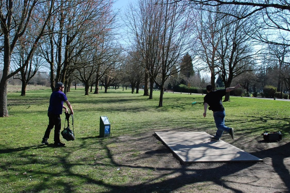 A disc golf player throws a green disc down the fairway from one of the tees at Blue Lake Regional Park disc golf course. Another person stands to the side and carries a bag of discs for the course.