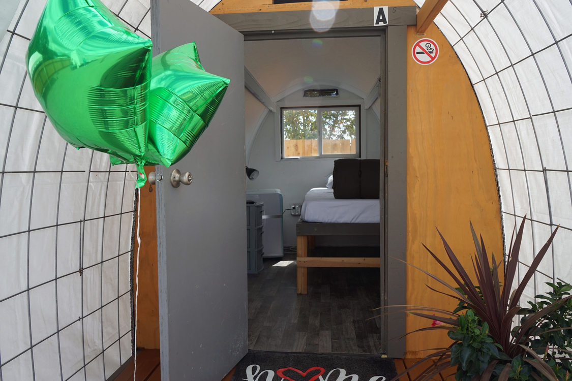Open door into a structure with a single bed and green balloons tied to the doorknob.