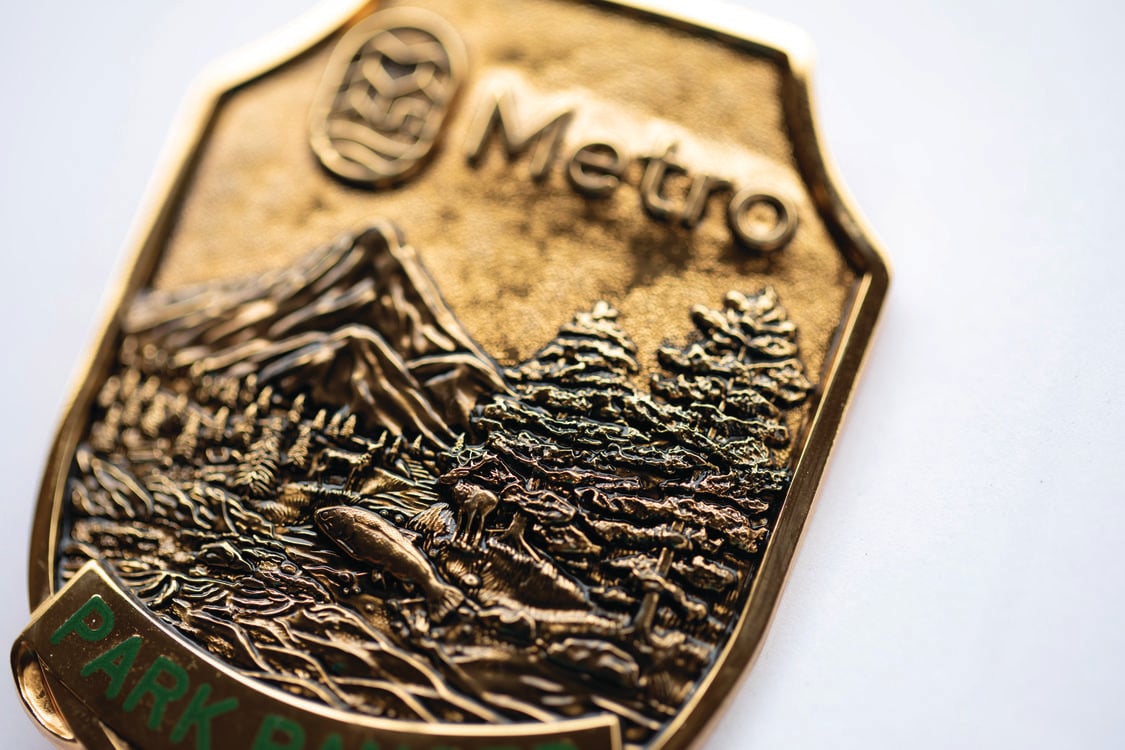 A close up of a bronze park ranger badge has details of mountains, trees and a river. It reads "Metro Park Ranger". 