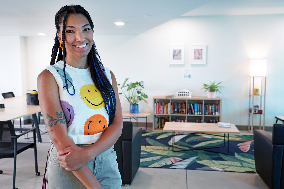 Young woman with long, multicolored braids smiling and standing in the community space of an apartment building.