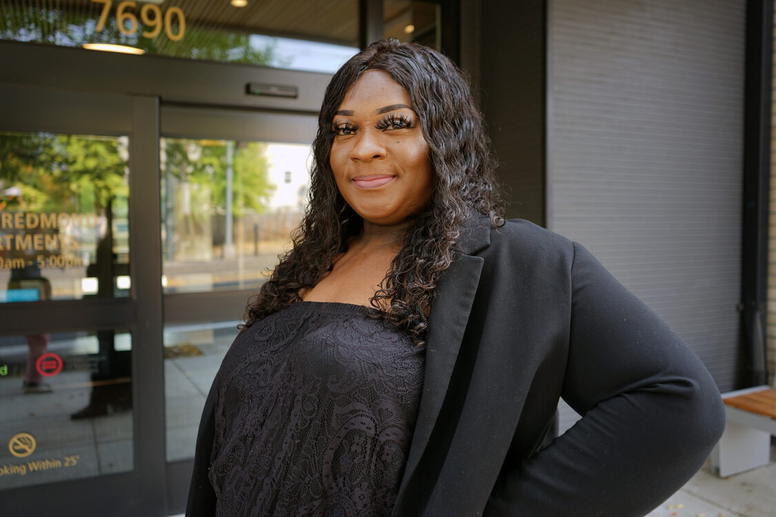 Woman dressed in all black standing in front of an apartment building smiling.