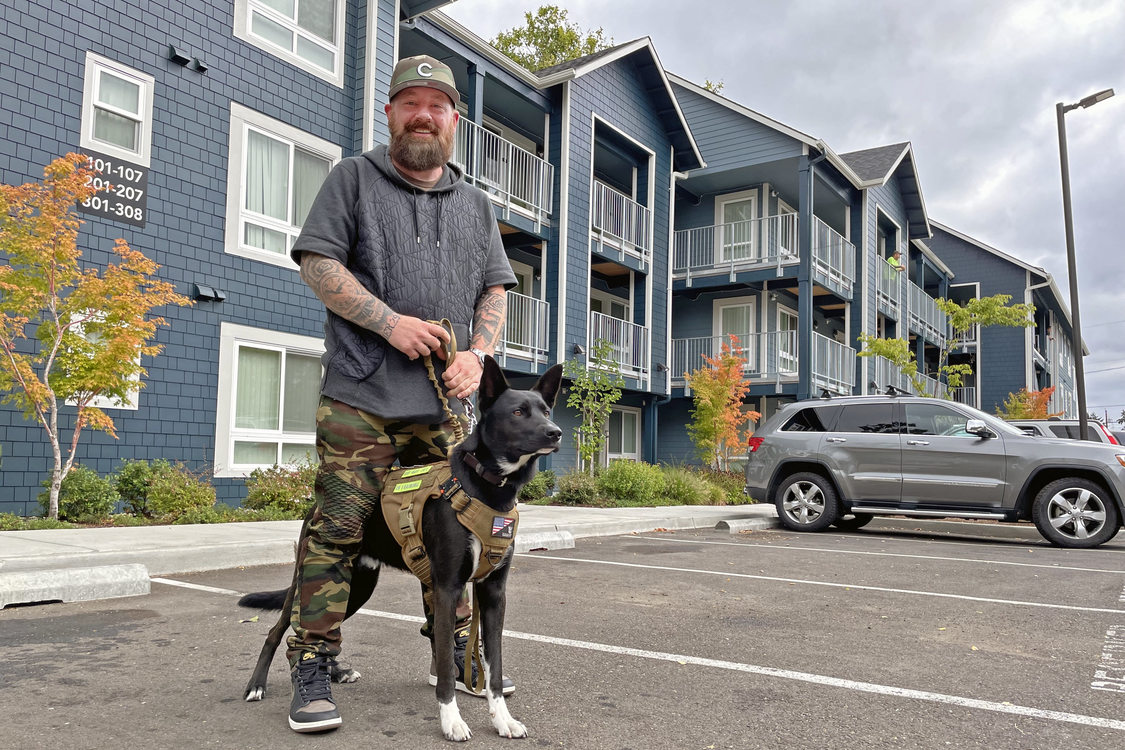 Man wearing a sweatshort and camo pants holding a dog leash and smiling, standing in front of an apartment building.