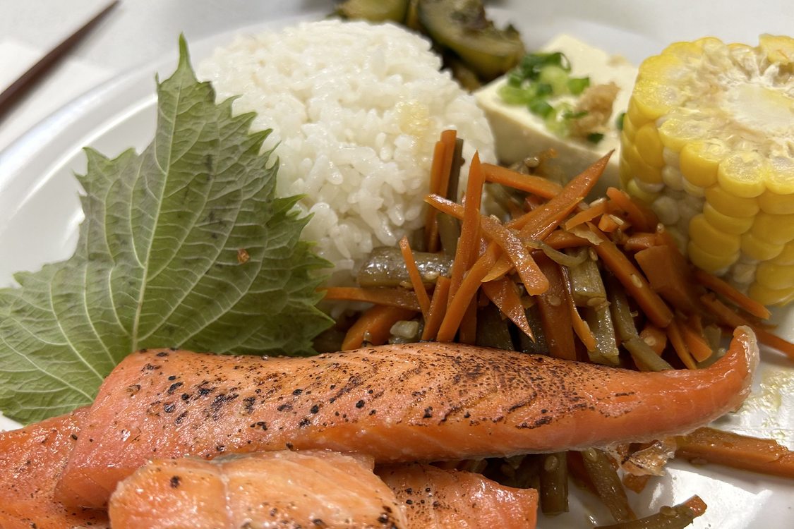 A white plate holds salmon, rice, corn, vegetables, tofu and soup.