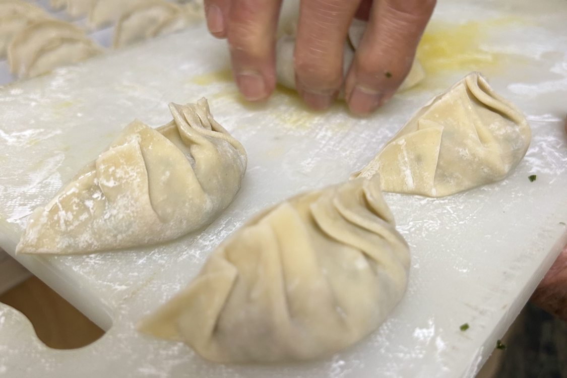 A hand paces a gyoza, a neatly folded Japanese dumpling, on a cutting board already holding three gyoza. In the background is a cutting board full of gyoza.