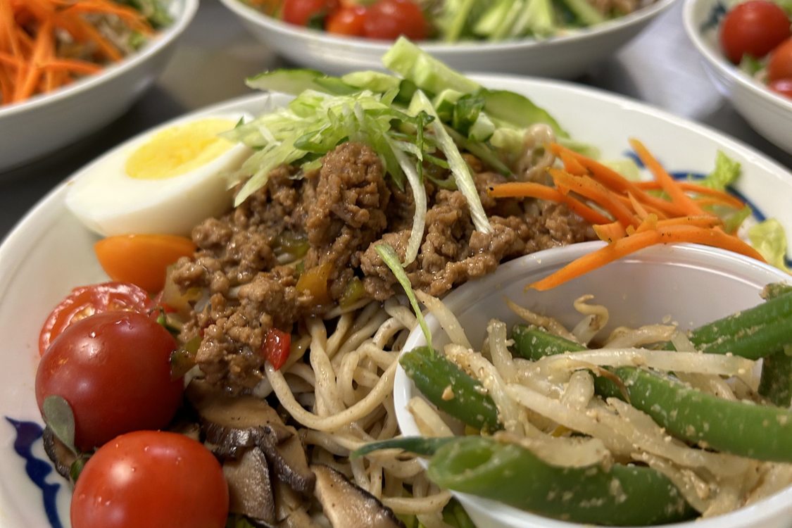 White plates with a swirly blue line holds noodles topped with meat, vegetables, cherry tomatoes, and half a hard-boiled egg.