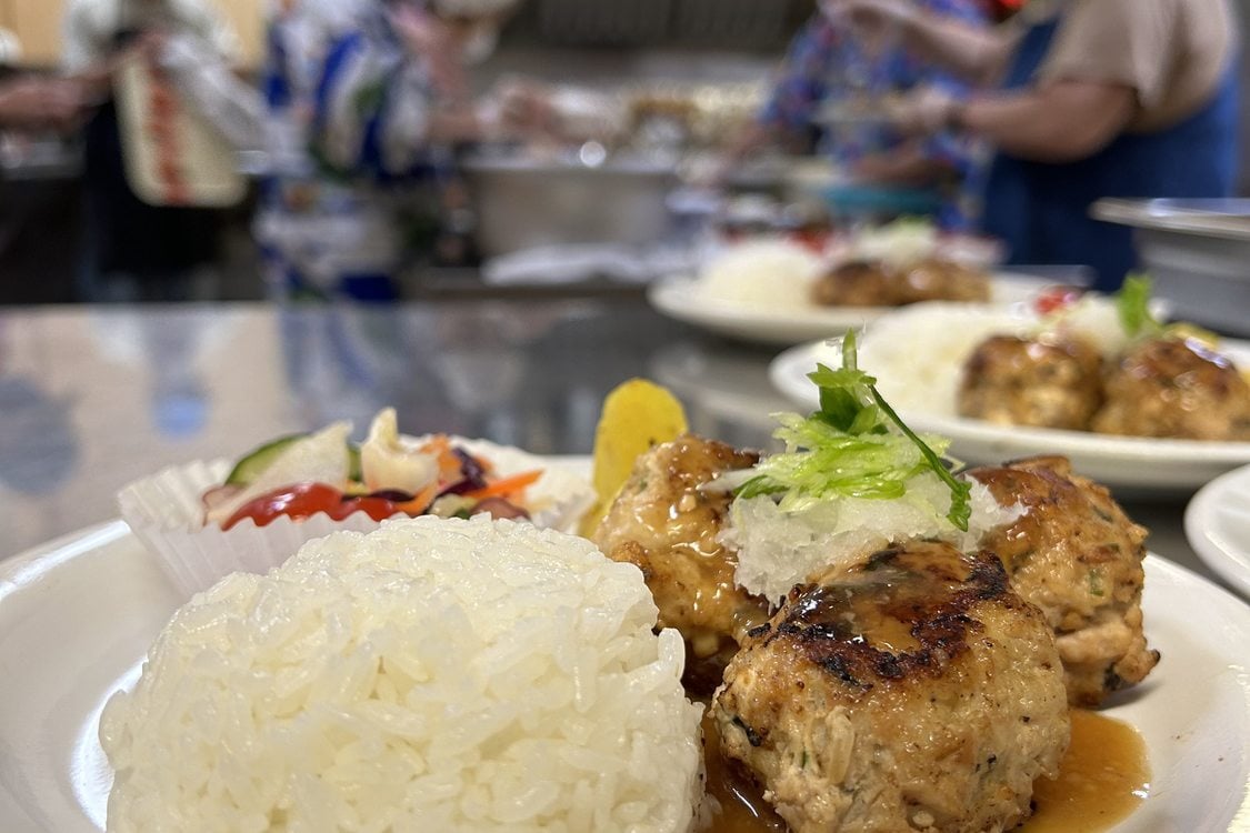 A white plate holds rice, three meatballs and some pickled vegetables. In the background, blurry figures stand in a stainless steel kitchen. 