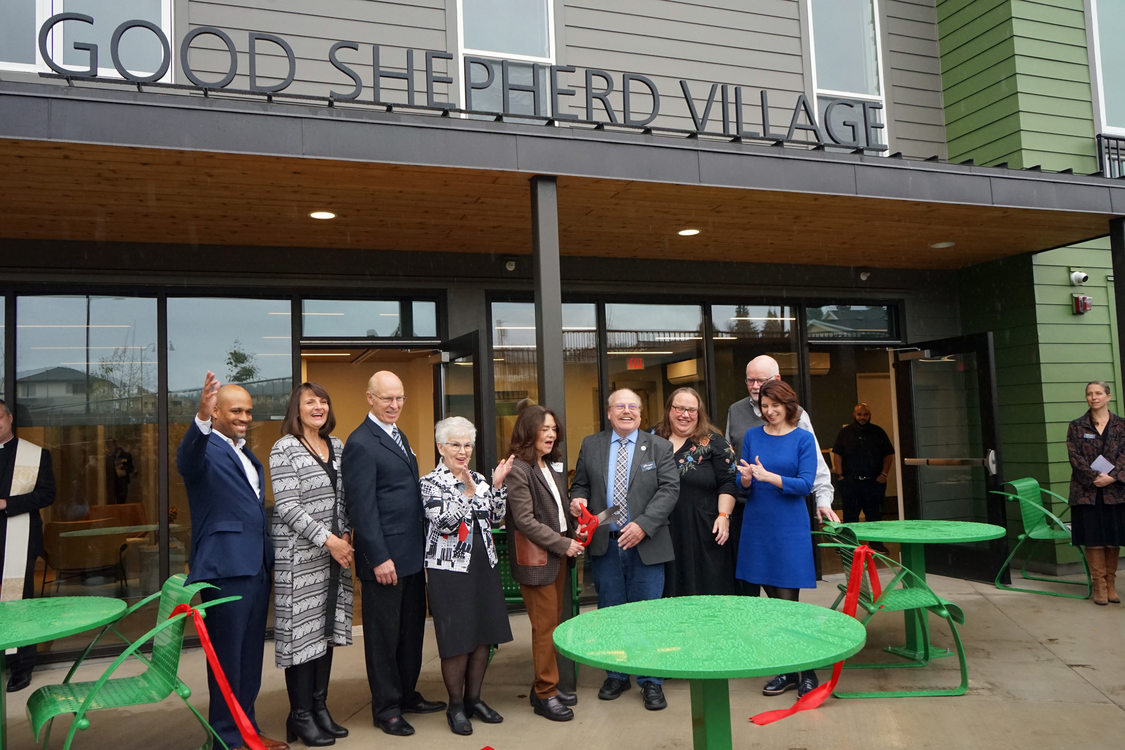 Red ribbon cutting with nine people in front of an apartment building with a sign reading "Good Shepherd Village."