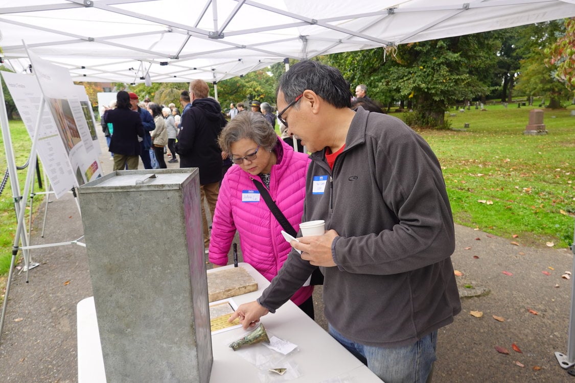 woman and man standing at a table under a canopy in a cemetery, looking at Chinese writing on a gravestone lying on the table