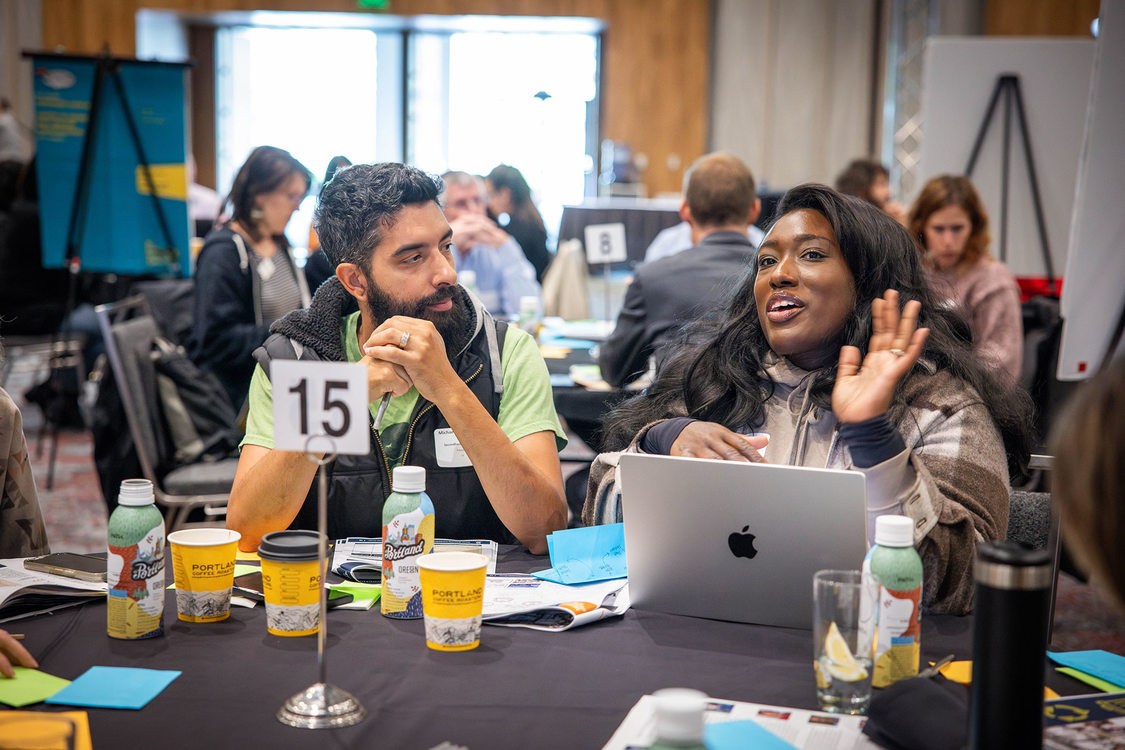 Focus on a man and woman sitting at a table, the woman is talking expressively, the man is watching the person speak. In the background other people are reading presentation materials.