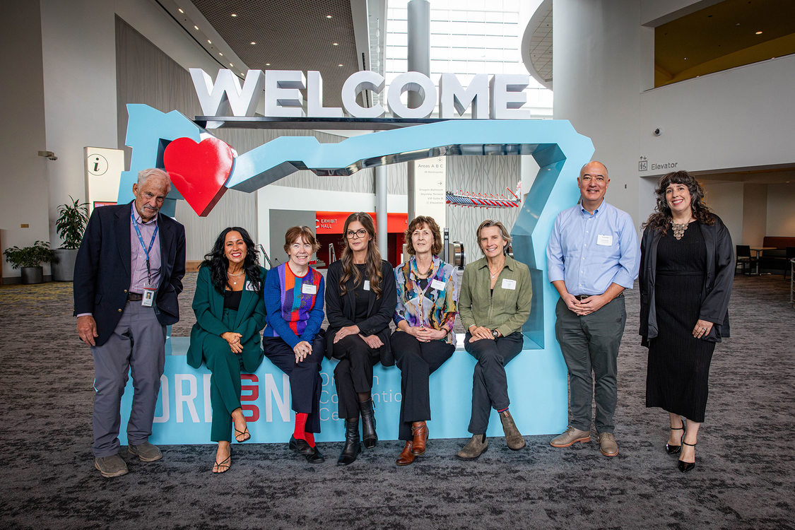 8 people are infront of a light blue cutout of Oregon, 5 are sitting in the cutout, 3 are standing