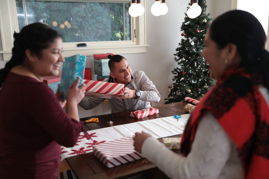Three people around a dining room table investigating wrapped presents with a Christmas tree in the background.