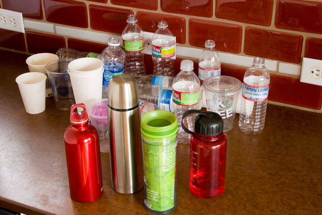 A counter top with reusable bottles and cups in front of plastic water bottles and throwaway coffee cups