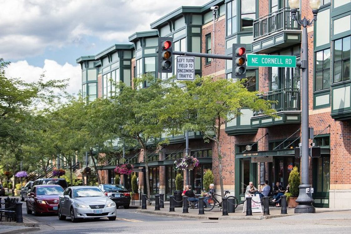 An active corner street in Orenco Station where cars are stopped at a stoplight. Trees line the sidewalk on both sides and people enjoy a restaurant with outdoor seating. 