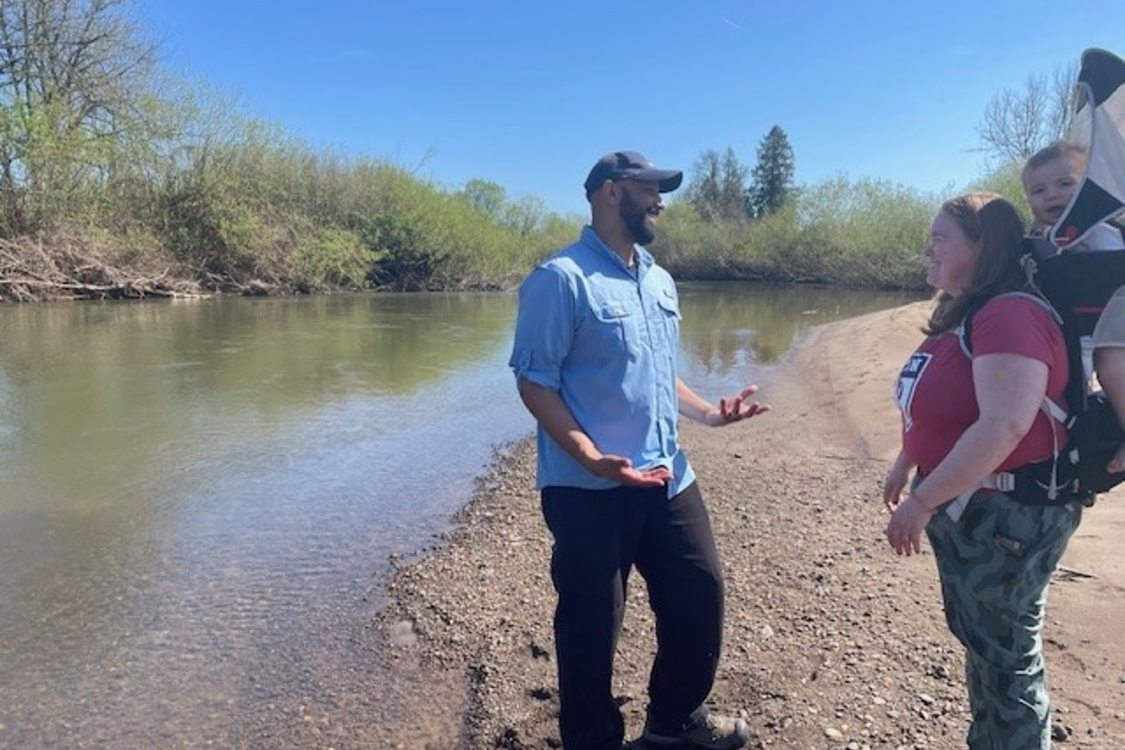 Metro Councilor Christine Lewis tours Metro’s Lower Pudding River Natural Area acquisition with Director Jon Blasher. She stands near a river's edge on a sunny day with a small child strapped to her back.
