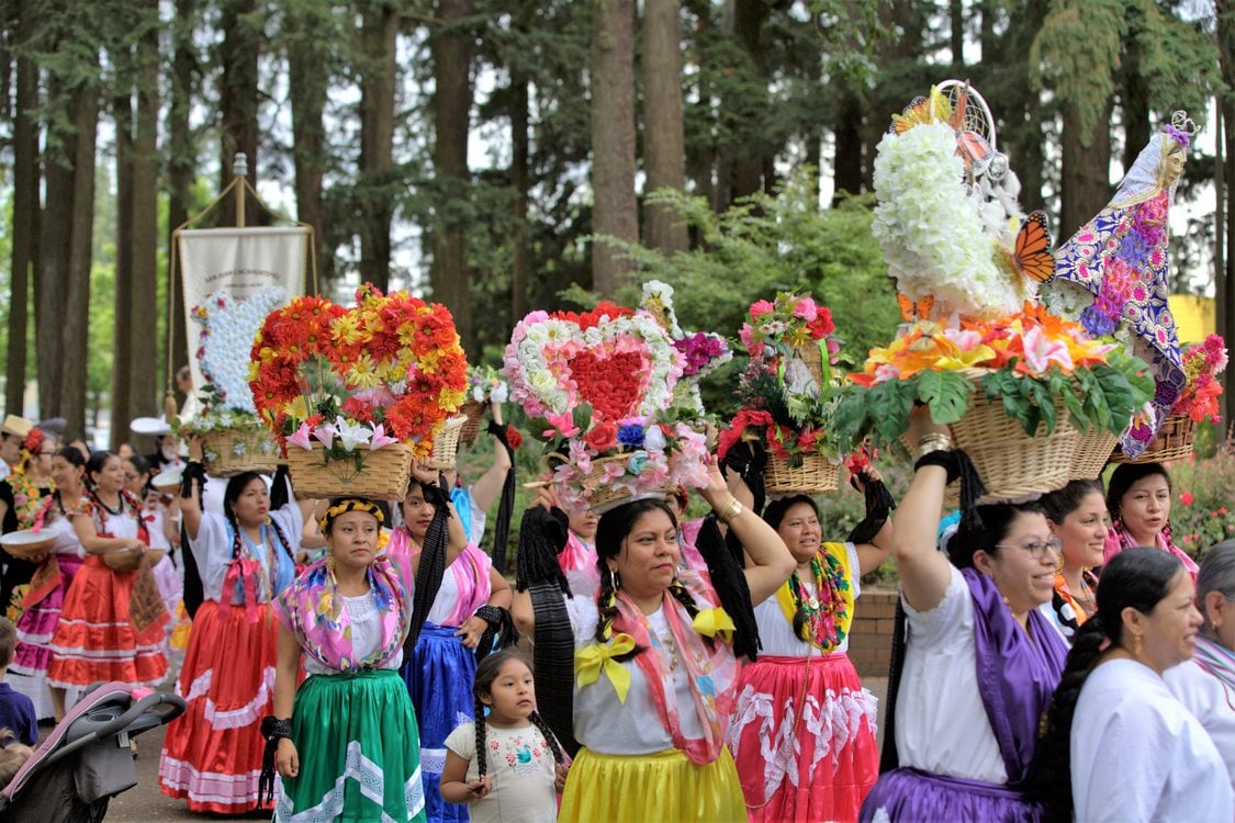 A large group of Latina women and girls of all ages wearing colorful, traditional clothing walking through a wooded park balancing ceremonial bouquets of flowers and art in baskets on their heads
