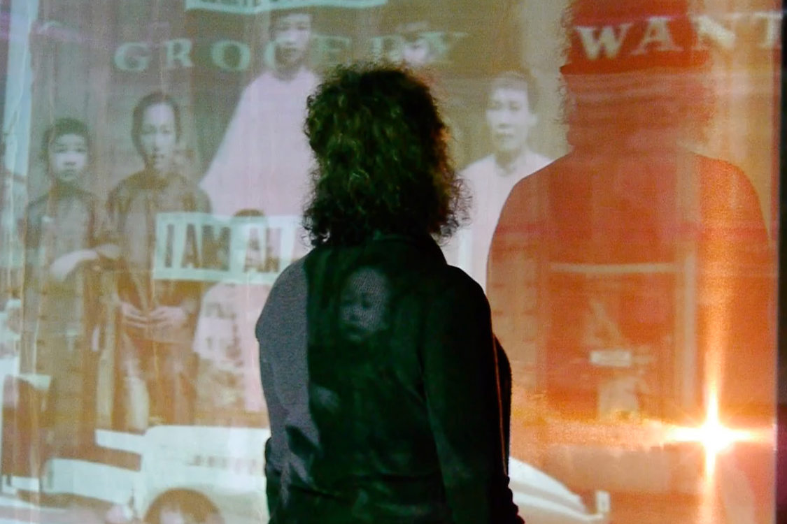A person stands facing a large projector screen that displays historical photos of Chinese immigrants from the early days of Portland's history