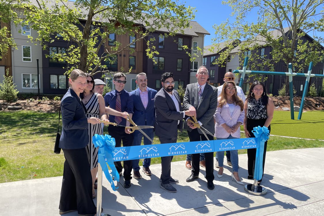 Ten people standing behind a ribbon that reads "Bienestar" in a grassy plaza surrounded by multifamily housing.