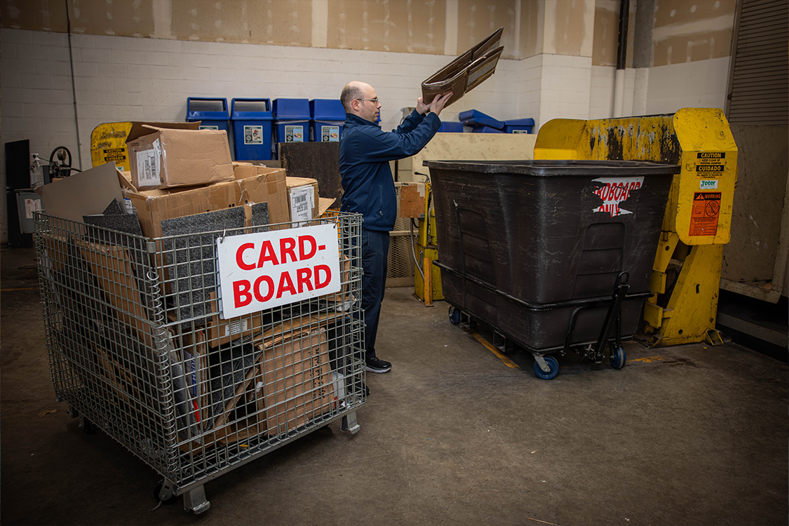 A man wearing a blue fleece jacket and glasses holds a piece of flattened cardboard high in the air before depositing it into a large black bin on wheels labeled "cardboard only" with other recycling bins and crates in the large room.