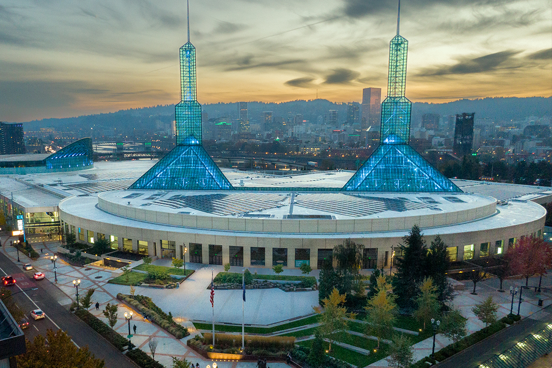 Oregon Convention Center at sunset, with the glass towers lit in blue and green lights and the rooftop solar panels in view.