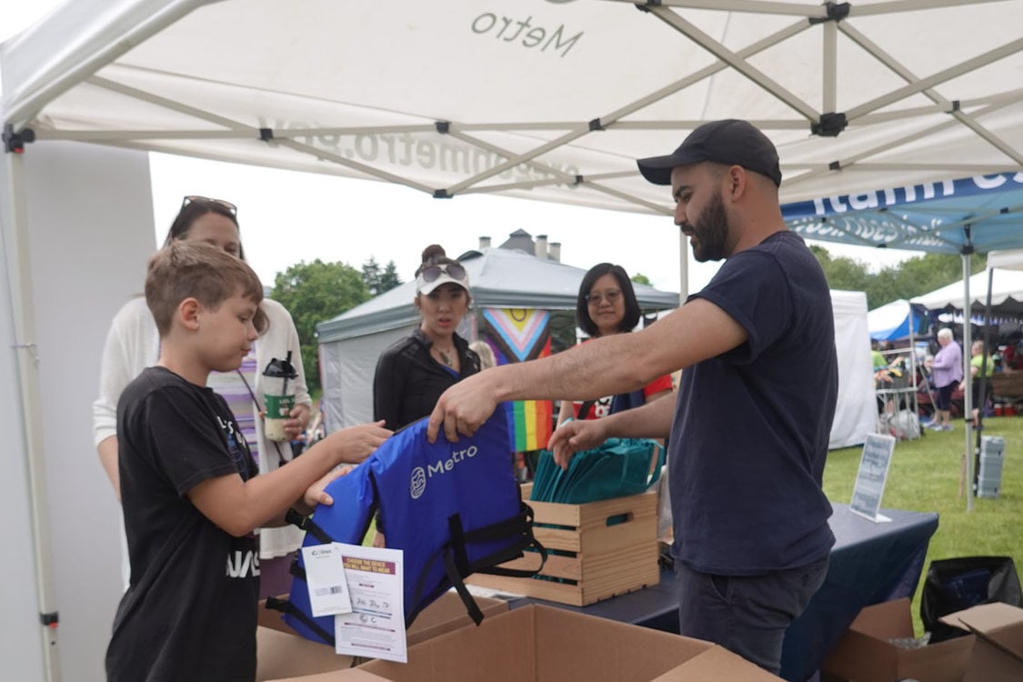 A Metro employee hands a child a Metro lifejacket at a festival while adult visitors look on