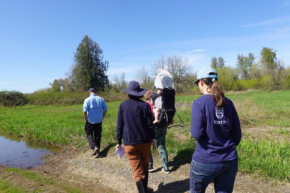 four adults, one carrying a baby in a backpack carrier, walk through a grassy field along a dirt road that submerges into a large puddle