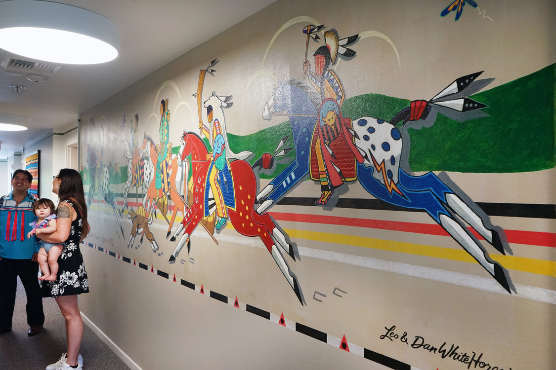 Large mural depicting Native Americans riding multicolored horses, with a man, woman and baby standing to the side chatting.