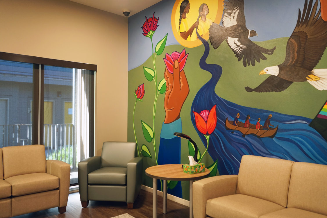 A community room with two tan sofas and a matching armchair around a small, round coffee table. On the wall is a large mural depicting various scenes: a mother and child surrounded by flowers, an eagle in flight, and a group in a canoe on a river.