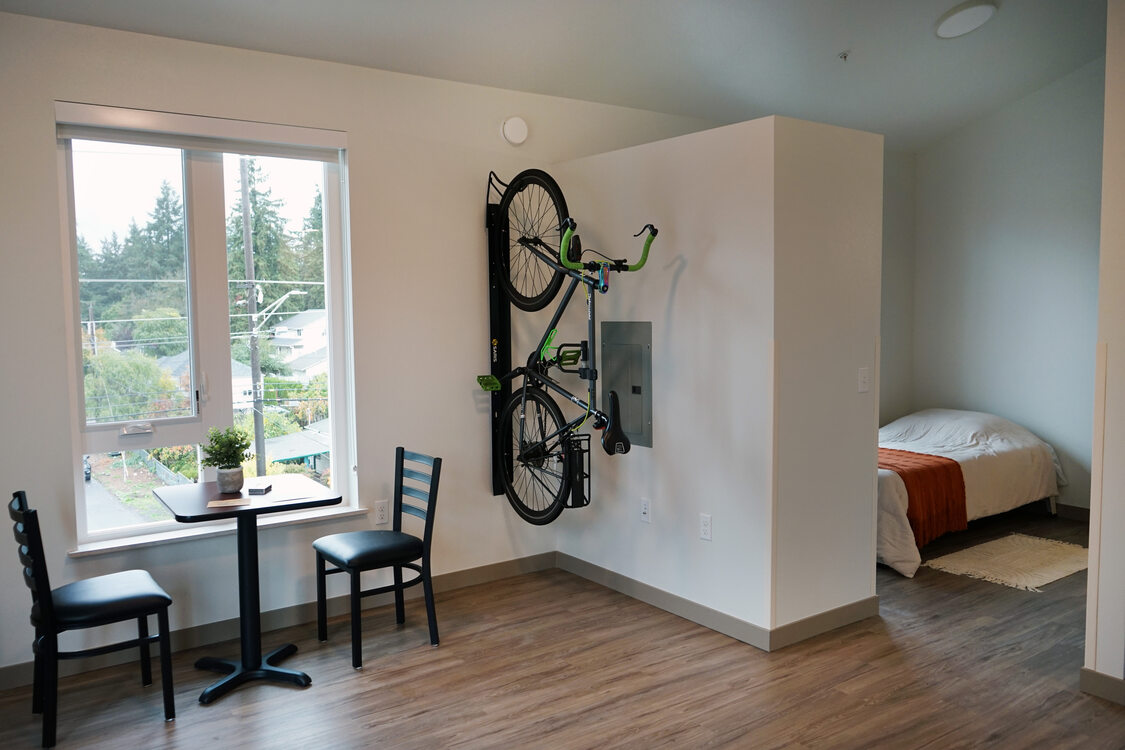 Studio apartment with large windows, table and chairs, bike rack and separate sleeping area.