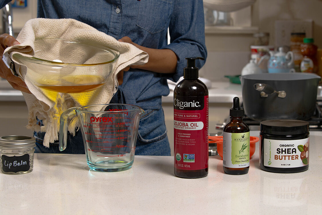 A person stands behind a counter about to pour melted oil into a container, making safe lip balm. The counter has a container for the balm, jojoba oil, eucalyptus essential oil and shea butter.