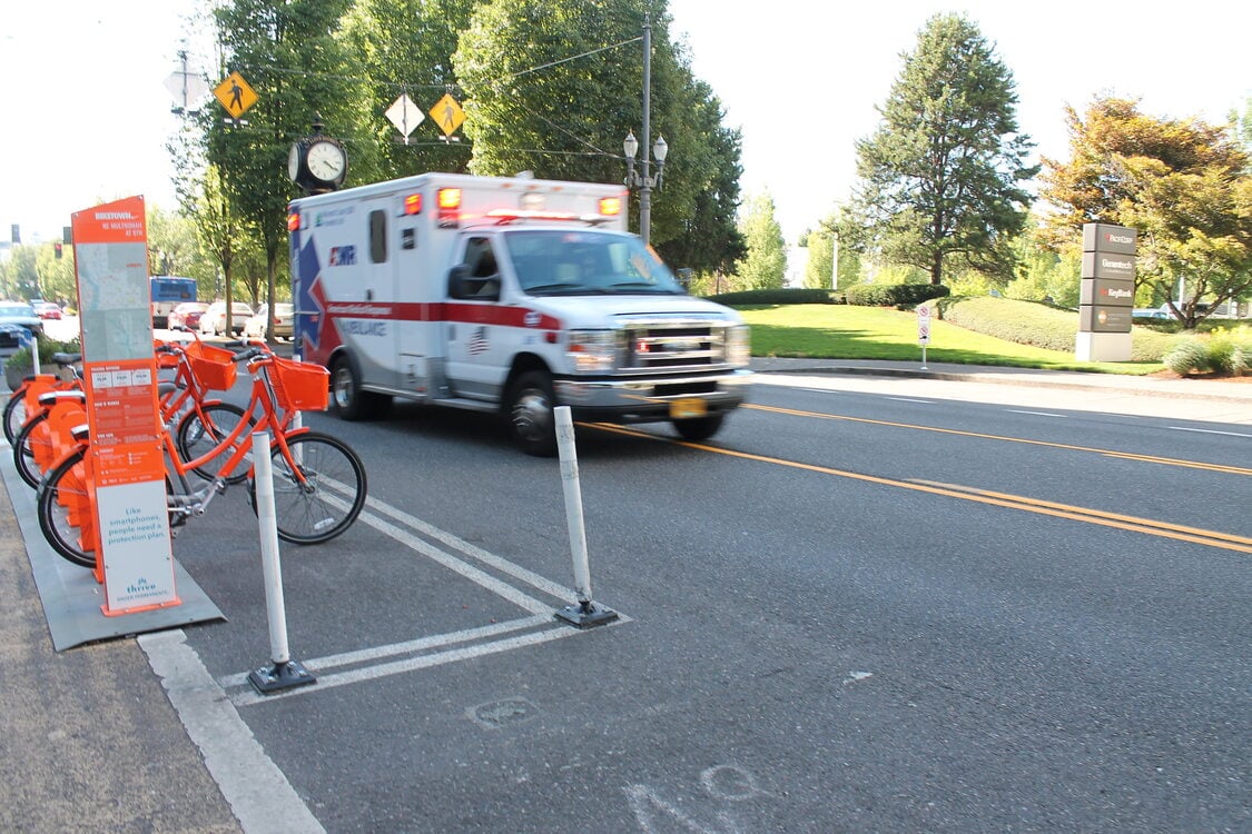 An ambulance with its emergency lights on drives along a road passing a Nike Biketown rack of bikes