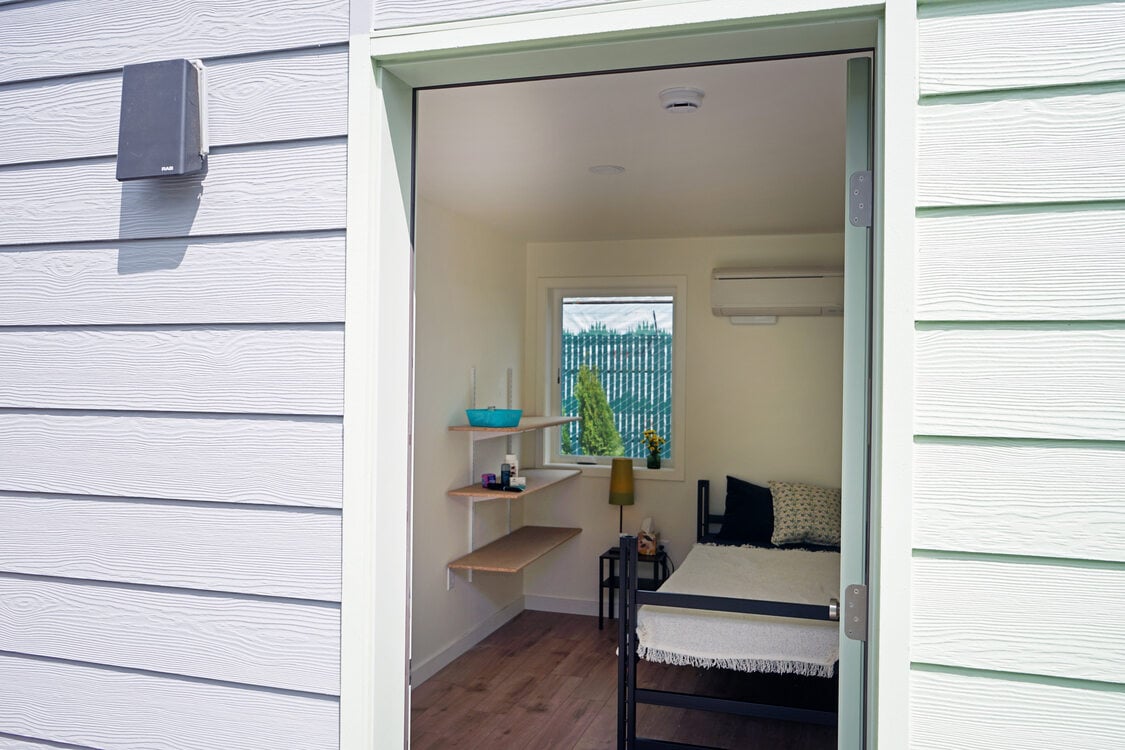View through an open door to the interior of a small living space with shelves, a window and a bed. 
