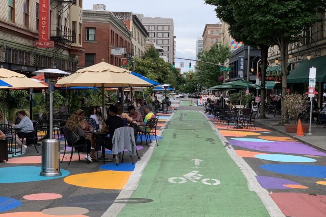 People sit and dine at tables covered by umbrellas on a colorfully painted plaza road that is blocked off from traffic and has a bike lane through it