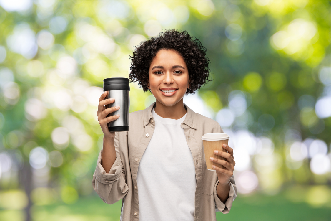 A woman holds two cups, one in each hand. The reusable cup she holds forward, the disposable cup she holds closer to her body, indicating that she is moving away from disposable cups