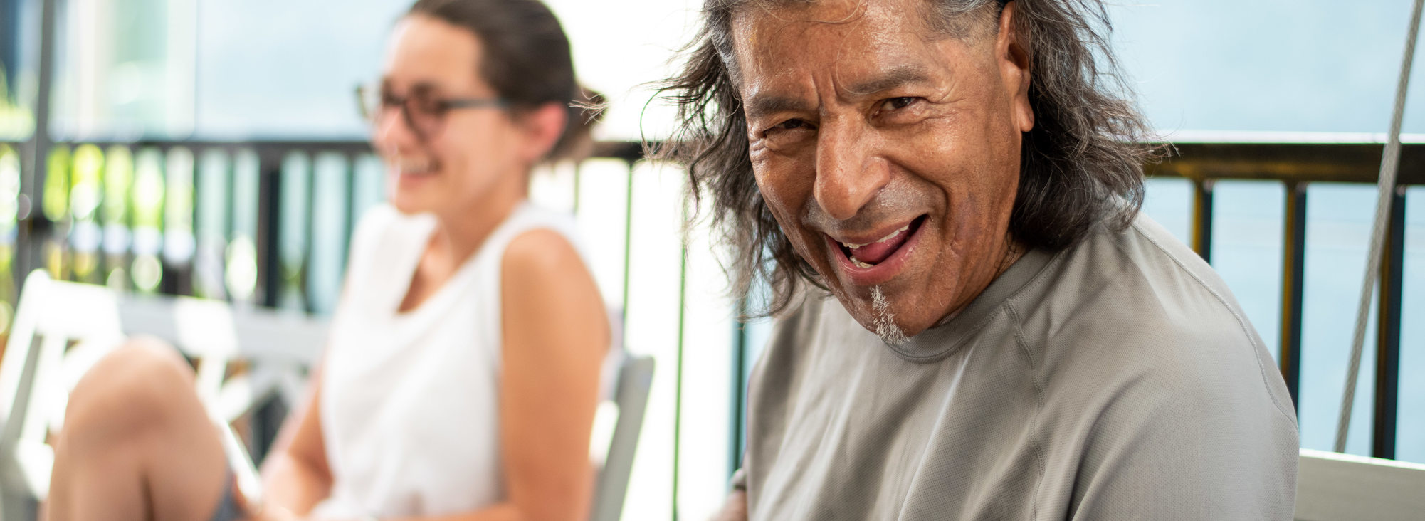 A smiling person with a tan shirt and sunglasses on their head and another smiling person blurred in the background