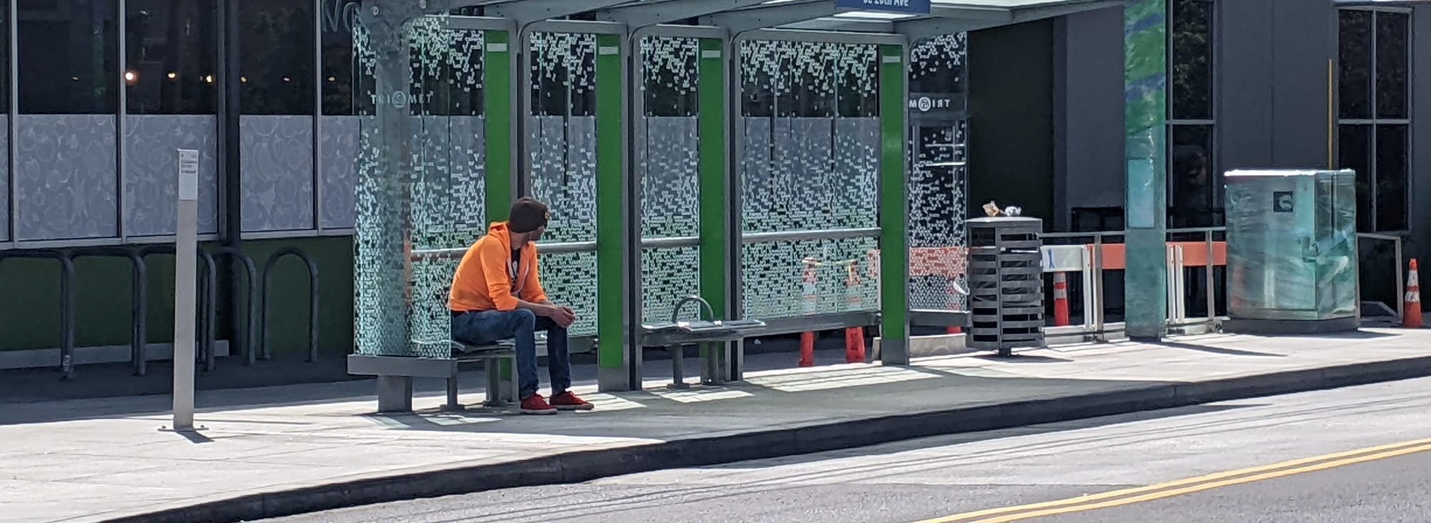 A man wearing a bright orange hoodie and red sneakers sits on a bench under a transit shelter on Southeast 20th Avenue in Portland