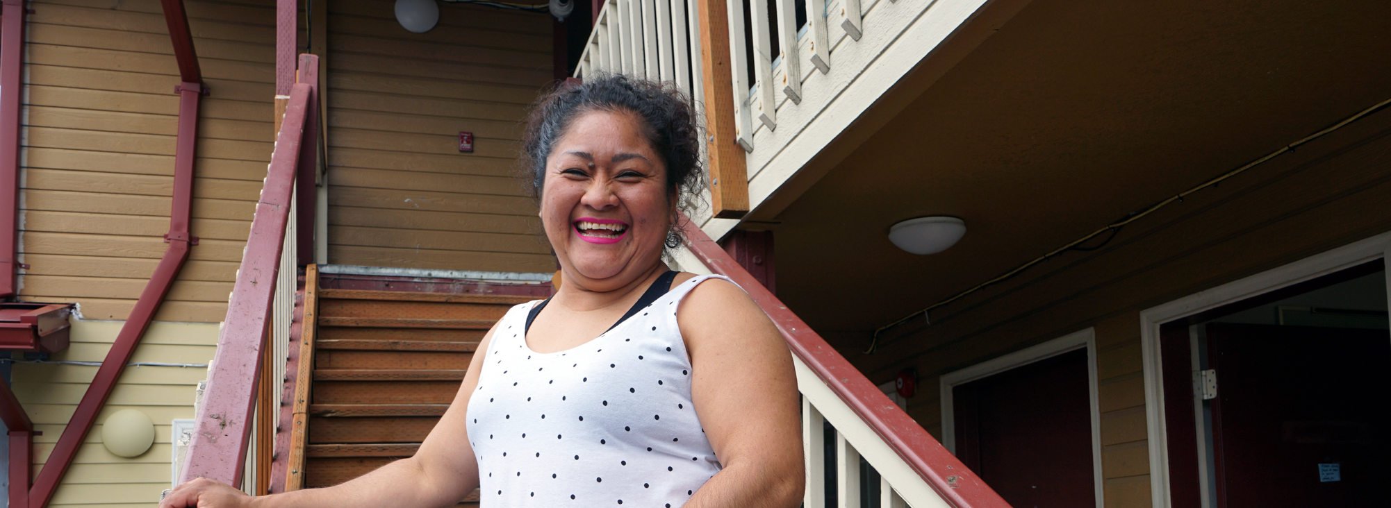 Latina woman in white polka dot tank top and black pants smiling and standing at foot of exterior stairs