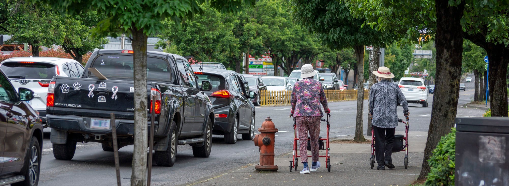 Two people with walkers walking on the sidewalk next to vehicles on the road.