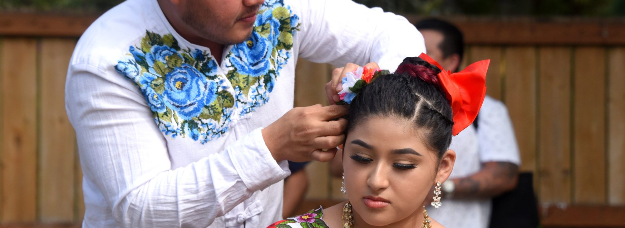 A person in a white shirt with blue flowers on the chest fixing a flower in the hair of another person wearing a multi-colored floral dress at the Mayan Culture and Tradition Fest