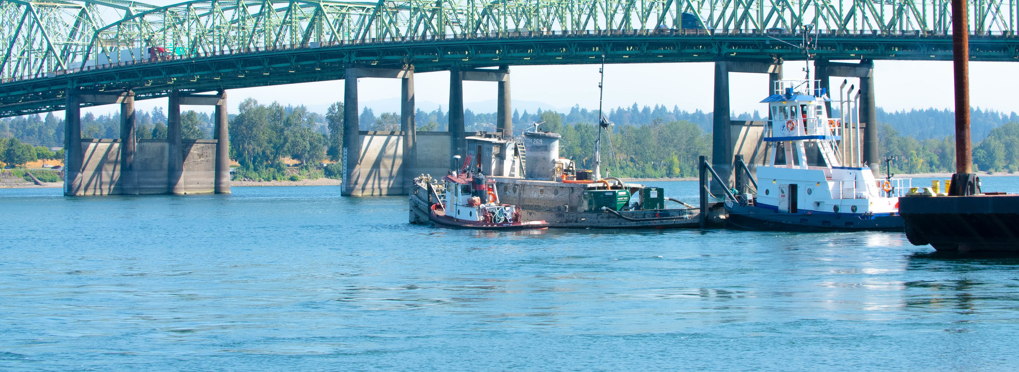 Two tugboats maneuver two derelict boats in the Columbia River, with the Interstate Bridge in the background.