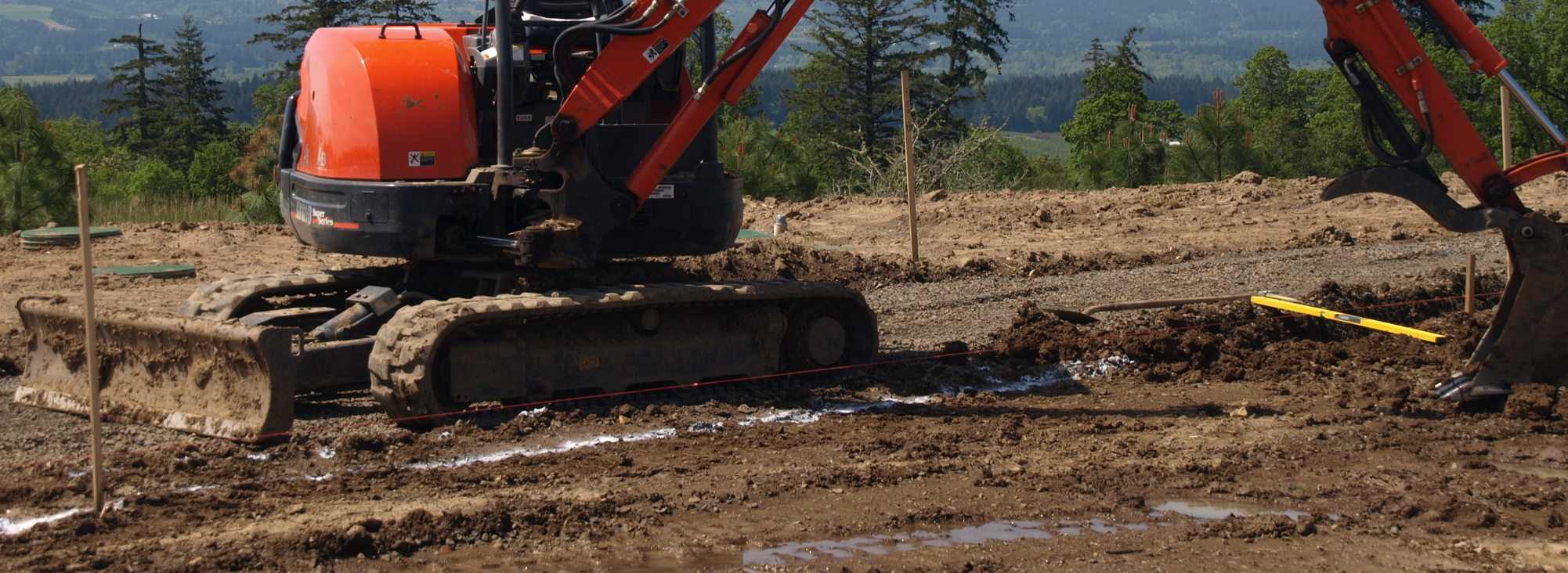 An orange construction digger with its bucket extended sits on a partially-leveled muddy parcel, on a sunny day with evergreen trees in the distance.