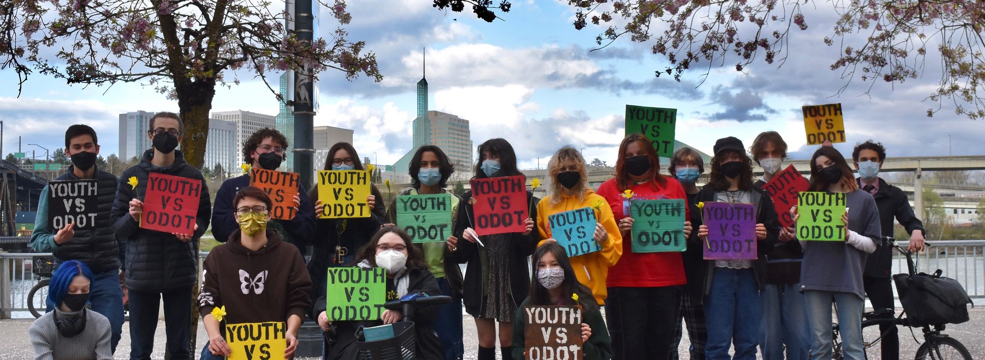 A group of youth at a climate justice rally hold signs that say "Youth Vs ODOT" by the waterfront in Portland, Oregon. Blooming, pink cherry blossoms frame the photo on each side. A bit of blue sky peaks through the clouds
