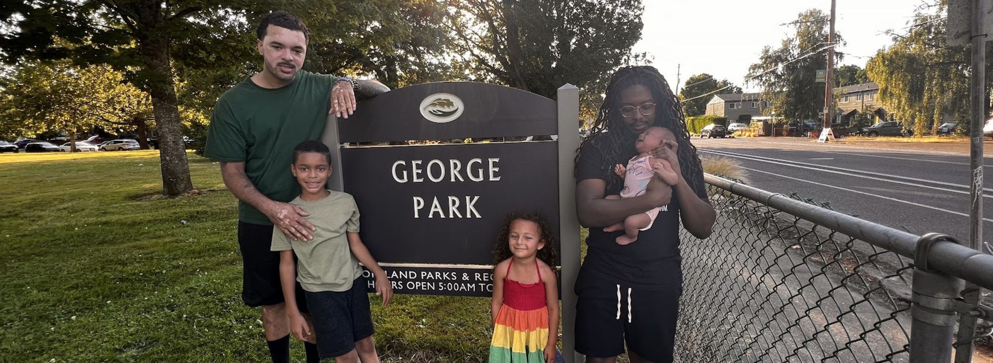 Mat Randol and his son pose on the left side of the George Park sign while Donovan Scribes holds his step daughter and baby to the right of the sign. The sun peaks through the trees in the background. 