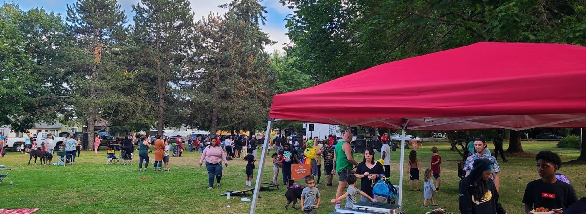 A wide view of the Kidz Outside Festival at George Park. In the foreground, people serve themselves food underneath a red tent. Trees and people line the background.