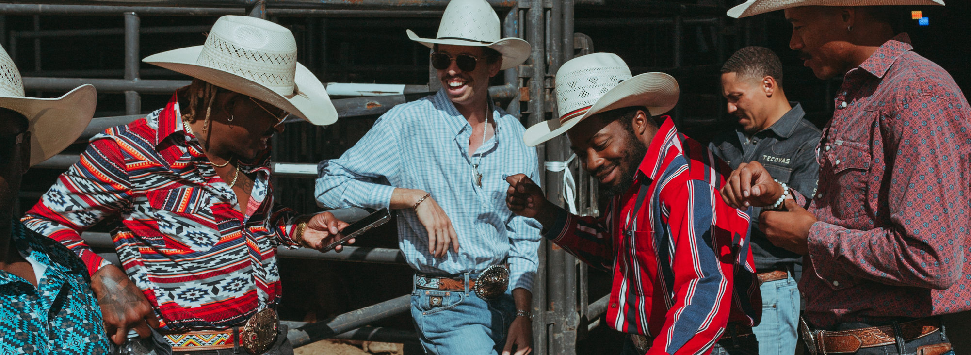 A group of rodeo participants wearing brightly-colored Western-style shirts and cowboy hats smile and dance near the metal animal corrals at Expo Center.