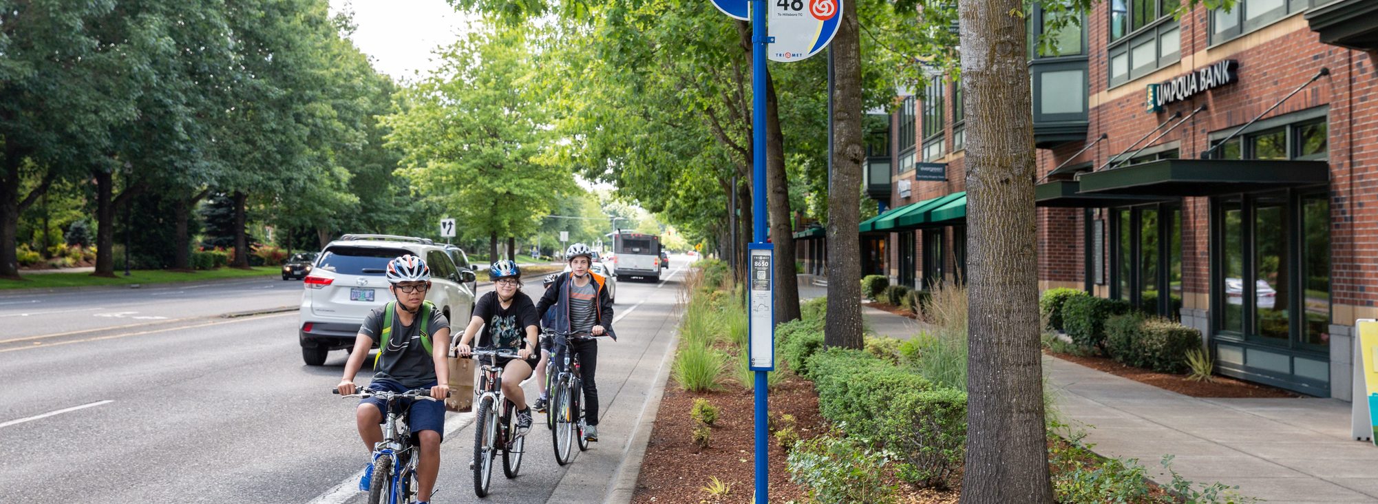 Three young people wearing helmets ride their bikes in a queue on the bike lane near the NE Cornell and Orenco Station bus stop next to an Umpqua Bank