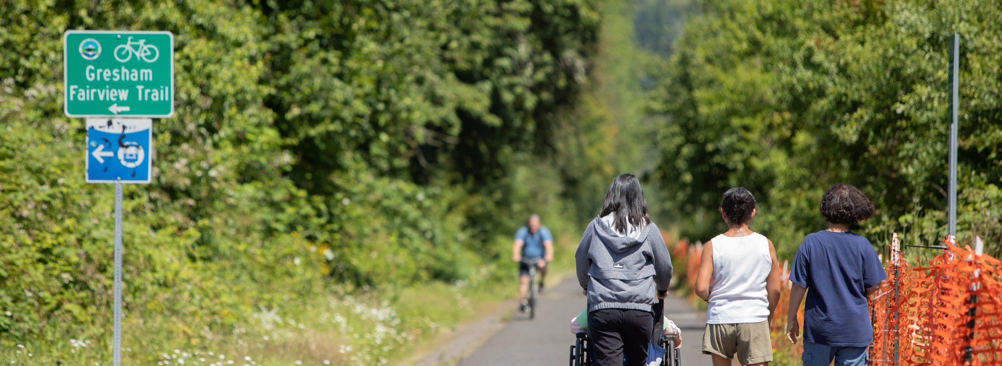 A group of three people walk along the Gresham Fairview Trail. One person pushes another person in a wheelchair. A person on a bike is approaching from further down the trail. Trees line both sides of the trail on a sunny day.